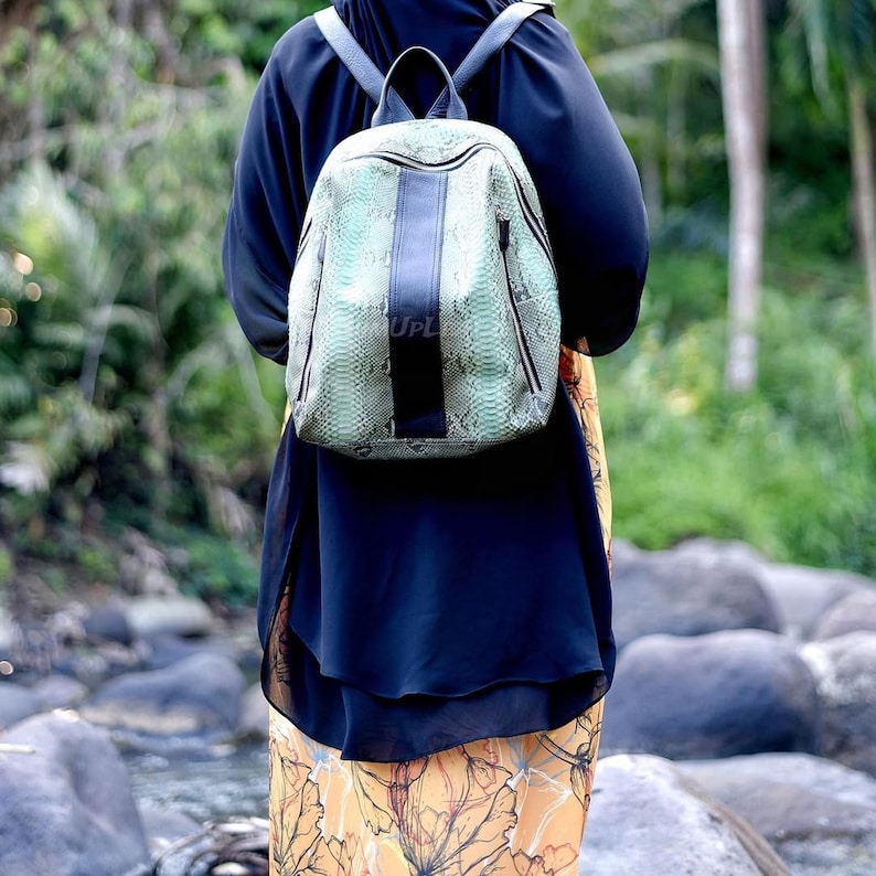 May include: A backpack featuring a light green and black snakeskin pattern with a solid black vertical center stripe. The bag has a top handle, two shoulder straps, and a zipper closure. The text UPL is visible on the center black stripe.