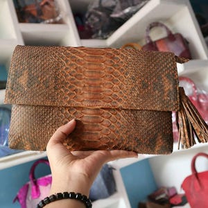 Könnte beinhalten: Eine braune Schlangenleder-Clutch mit Klappe und Quaste. Die Clutch wird in einer Hand gehalten, wodurch die Textur und das Muster des Schlangenleders hervorgehoben werden. Weitere Taschen sind im Hintergrund zu sehen.
