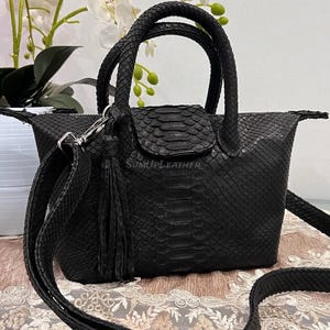 May include: Black snakeskin-effect handbag with a top handle and a detachable shoulder strap. The bag has a flap closure and a decorative tassel. The bag is in front of a white vase with flowers.