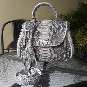 May include: A grey and white snakeskin-patterned handbag with a top handle and a detachable shoulder strap. The bag features a flap closure with a silver-tone clasp, a tassel, and a zip detail. The bag is on a glass table.