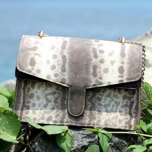 Peut inclure: Sac à main rectangulaire à rabat, avec un motif peau de serpent dans les tons gris et crème. Le sac a un accent noir et une bandoulière chaîne dorée. Il repose sur un rocher au milieu d'un feuillage vert, avec un arrière-plan bleu de l'océan.