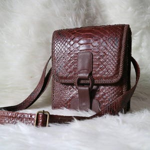 Könnte beinhalten: Braune Leder-Umhängetasche mit strukturierter, schuppenartiger Musterung. Die Tasche hat einen Klappenverschluss mit Schnalle, einen langen, verstellbaren Riemen und einen geflochtenen Rand. Die Tasche ist vor einem weißen, flauschigen Hintergrund platziert.