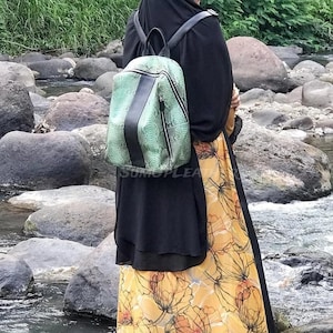 May include: A backpack featuring a light green snakeskin-textured pattern with a solid black vertical center stripe and black shoulder straps. The bag has a zippered main compartment and is worn over a black garment and a yellow floral print skirt.
