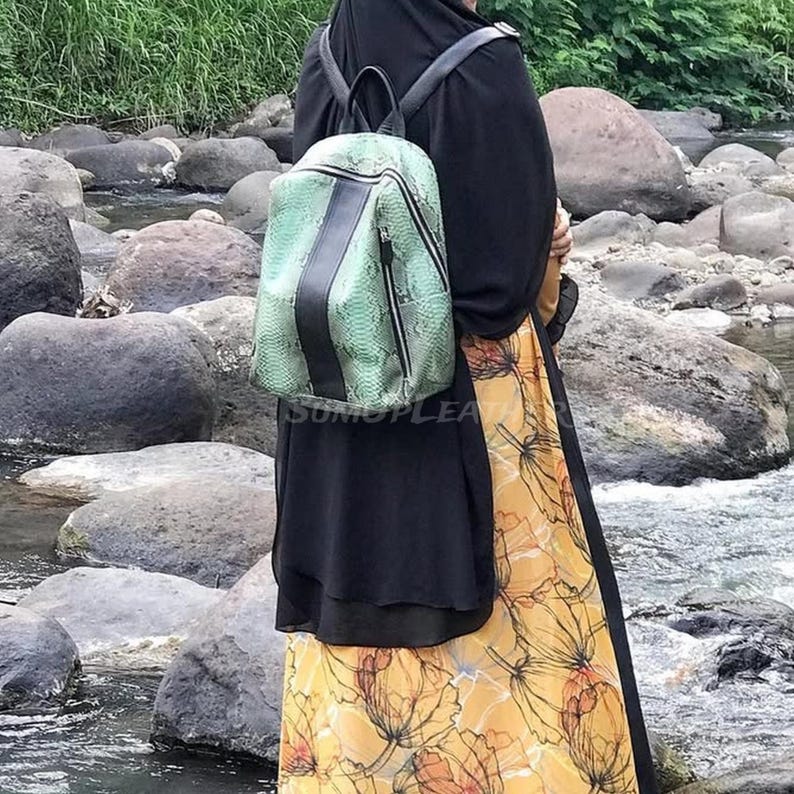 May include: A backpack featuring a light green snakeskin-textured pattern with a solid black vertical center stripe and black shoulder straps. The bag has a zippered main compartment and is worn over a black garment and a yellow floral print skirt.