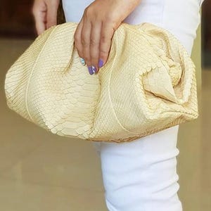 Peut inclure: Une pochette de couleur crème avec un motif texturé en peau de serpent. Le sac présente une silhouette souple, arrondie et froncée avec une fermeture de type cadre. La surface extérieure affiche une texture d'écailles répétitive et détaillée.