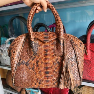 Op de afbeelding: Een bruine en zwarte handtas van slangenleer met een gevlochten handvat en een kwast. De tas heeft een afgeronde vorm en een ritssluiting. Andere handtassen zijn zichtbaar op de achtergrond.