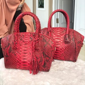 May include: Two red python skin handbags with matching handles. The bags have a textured, scale-like pattern and a tassel detail. One bag has a zipper closure. The bags are on a white surface.