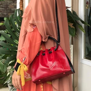 Könnte beinhalten: Eine rote Handtasche mit strukturierter Oberfläche und schwarzen Akzenten. Die Tasche hat einen Kordelzugverschluss und einen schwarzen Griff. Die Tasche wird über der Schulter getragen. Der Markenname "SUMUPLEATHER" ist sichtbar.