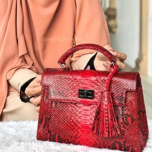 May include: A red handbag with a snakeskin pattern, a top handle, and a decorative tassel. The bag has a rectangular shape with a flap closure and a silver-coloured clasp. The text "SUMUPLEATHER" is visible on the top left.
