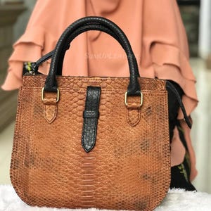 Könnte beinhalten: Eine braune Lederhandtasche mit Schlangenmuster. Die Tasche hat schwarze Griffe und einen schwarzen Riemenverschluss. Goldfarbene Hardware-Akzente zieren die Tasche. Der Text "SUMUPLEATHER" ist auf der Tasche sichtbar.