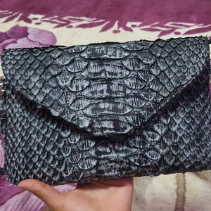 May include: A rectangular envelope-style clutch bag featuring a textured, scale-like pattern in shades of dark charcoal gray and black. The surface has a distinct, layered appearance with a pointed flap closure that covers the front of the bag.