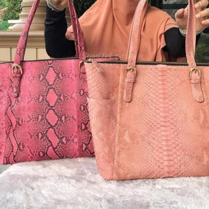 Könnte beinhalten: Zwei Ledertaschen in Rosatönen. Die Tasche links ist ein kräftiges Pink mit Schlangenmuster, die Tasche rechts ist ein helleres, pfirsichfarbenes Pink. Beide Taschen haben lange Henkel und goldfarbene Beschläge. Geeignet für den täglichen Gebrauch.