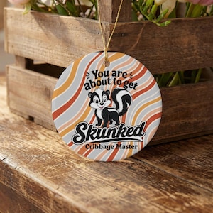 May include: A round ceramic ornament featuring a cartoon skunk illustration centered on a background of wavy orange, white, and grey stripes. The text reads: You are about to get Skunked Cribbage Master. A gold string is attached to the top.