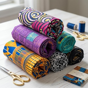 May include: A stack of six fabric rolls featuring diverse patterns in vibrant colors including yellow, purple, pink, teal, black, and white. The rolls are arranged on a surface with gold scissors and a small label reading Curated African Wax Prints.