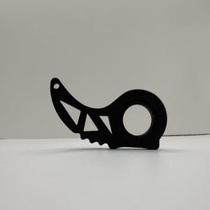 May include: A black, abstract-shaped object with a circular opening and geometric cutouts. The design includes a small hole at the top and a jagged edge on the bottom. The object is set against a plain white background.
