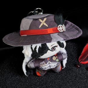 May include: A plush doll with a hat and a keychain. The hat is gray with a red band and a feather. The doll has a detailed face and a patterned outfit. The background is black with red accents.