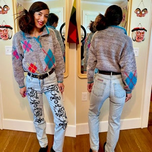 May include: A gray and white sweater with a diamond pattern in blue, pink, and yellow. The sweater has a mock neck and a button closure. The person is wearing light blue jeans with a black floral design.