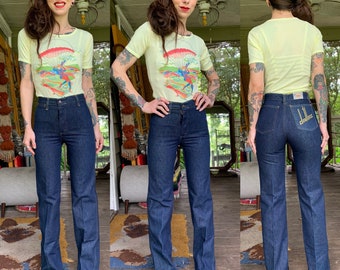 Può includere: Una donna indossa una maglietta giallo chiaro con una grafica di un paracadutista. Indossa anche jeans blu denim a vita alta con la scritta "DRIVES" ricamata sulla tasca posteriore.