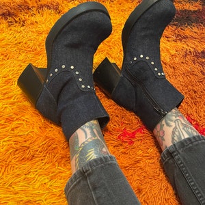 May include: A pair of dark blue denim ankle boots with silver studs on a thick platform sole. The boots are worn by a person with tattoos on their legs and are on a shaggy orange rug.