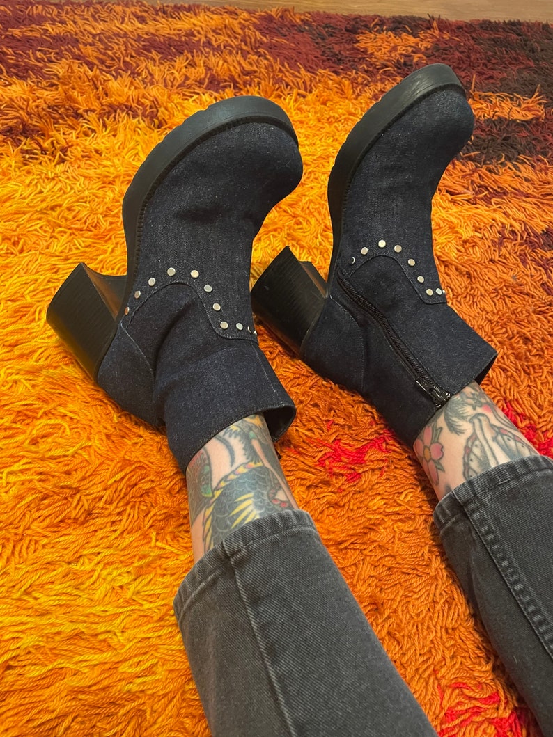 May include: A pair of dark blue denim ankle boots with silver studs on a thick platform sole. The boots are worn by a person with tattoos on their legs and are on a shaggy orange rug.