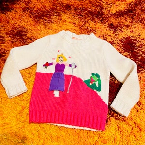 Può includere: Maglione di lana bianco e rosa con un'applicazione di maiale rosa e viola e una rana verde. Il maiale tiene un microfono e la rana tiene un fiore rosso.