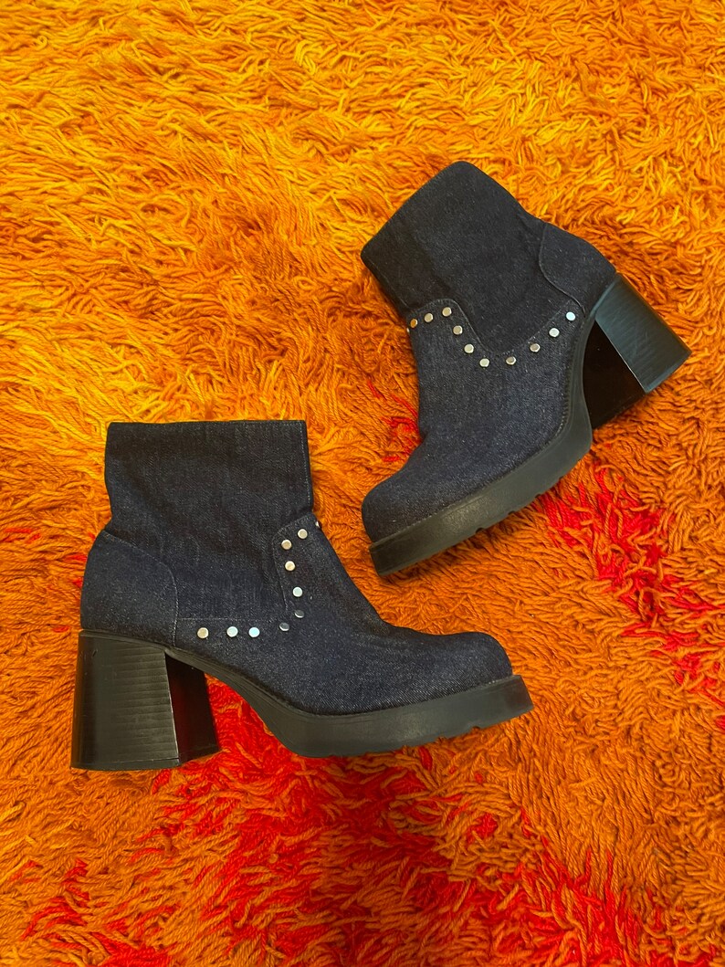 May include: A pair of denim ankle boots with silver studs and chunky heels. The boots are on a fuzzy orange rug.
