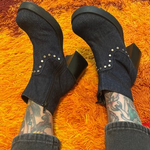 May include: A pair of dark blue denim ankle boots with silver studs. The boots have a chunky heel and a platform sole. The boots are worn by a person with tattoos on their legs.
