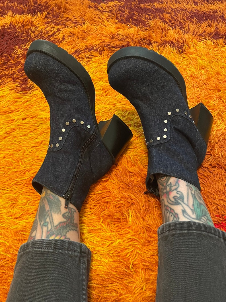 May include: A pair of dark blue denim ankle boots with silver studs. The boots have a chunky heel and a platform sole. The boots are worn by a person with tattoos on their legs.