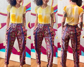 May include: A person wearing a yellow tie-dye t-shirt and colorful patterned pants. The pants have a geometric design with red, blue, green, and yellow colors.
