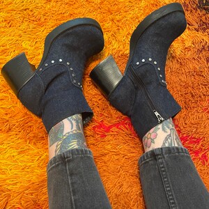 May include: A pair of dark blue denim platform boots with silver studs. The boots are worn by a person with tattoos on their legs and are on a shaggy orange rug.