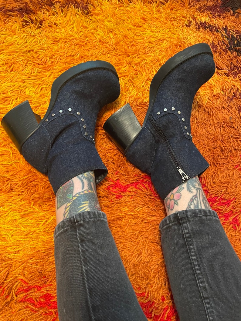 May include: A pair of dark blue denim platform boots with silver studs. The boots are worn by a person with tattoos on their legs and are on a shaggy orange rug.