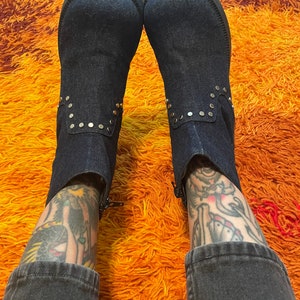 May include: A pair of dark blue denim boots with gold studs. The boots are worn by a person with tattoos on their legs. The boots are on a shaggy orange and brown rug.