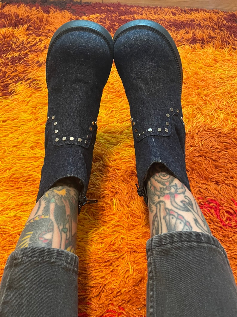 May include: A pair of dark blue denim boots with gold studs. The boots are worn by a person with tattoos on their legs. The boots are on a shaggy orange and brown rug.