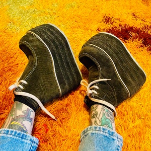 May include: A pair of olive green corduroy platform shoes with white laces and soles. The shoes have a thick, ridged platform and a gold trim. The shoes are on a bright yellow and orange shag carpet.