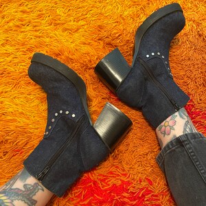 May include: A pair of dark blue denim ankle boots with silver studs and chunky black heels. The boots are on a shaggy orange and red rug.
