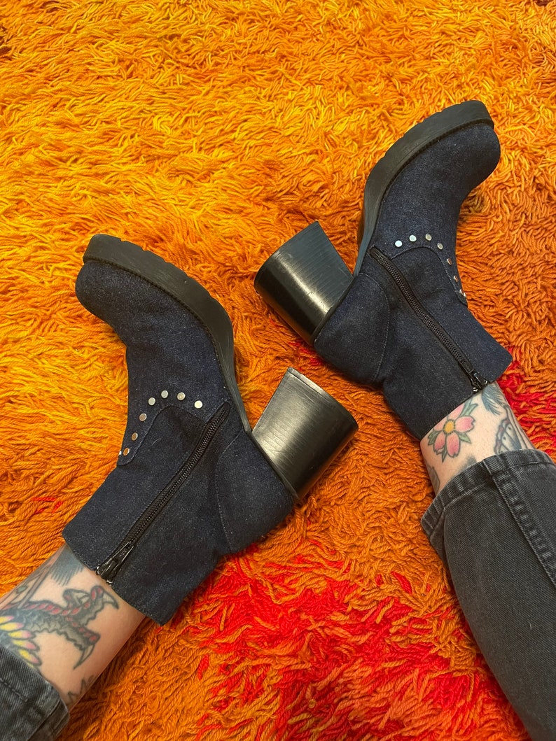 May include: A pair of dark blue denim ankle boots with silver studs and chunky black heels. The boots are on a shaggy orange and red rug.
