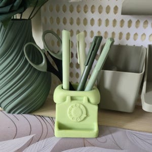 May include: A light green telephone-shaped desk tidy holding pens. The organiser has a rotary dial design and a small tray. Other items include a green vase with a plant and a pair of scissors.