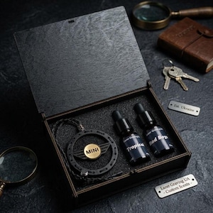May include: A dark wooden box, open to reveal a black pendant with a gold "MINI" emblem, and two small black bottles labelled "Tropical" and "Bad Boy". The box has a black interior.