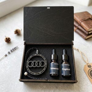 以下が含まれることがあります： 黒い木製の箱で、ヒンジ付きの蓋があり、車のエンブレムペンダント、2つの小さなガラス瓶、黒い内装が入っています。「Tobacco」と「Tropical」とラベルされた瓶。ペンダントはアウディのロゴが入った円形デザインです。