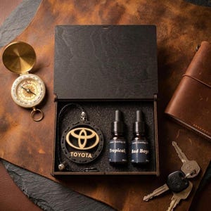 May include: A dark wooden box set featuring a Toyota logo pendant, two small bottles, and a leather cord. A vintage compass and a leather notebook are also present. The bottles are labelled "Tropical" and "Bad Boys".