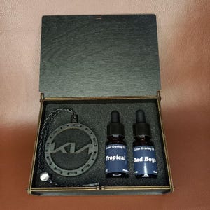 以下が含まれることがあります： 暗い木製の箱には、車の芳香剤、2つの小さなボトル、装飾的なKiaのロゴが入っています。ボトルには黒いスポイトと、「Tropical」と「Bad Boys」のテキストが入った青いラベルが付いています。芳香剤は、Kiaのロゴが付いた丸い形をしています。
