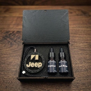 Puede incluir: Una caja de madera negra con tapa con bisagras, que contiene un colgante con el logotipo de Jeep, dos pequeños frascos cuentagotas y un cordón de cuero negro. Las botellas están etiquetadas como "Tropical" y "Bad Boys".