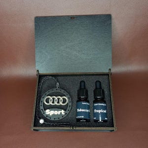 May include: A dark wooden box with a hinged lid, containing a car air freshener and two small glass bottles. The air freshener features the Audi logo and the word "Sport". The bottles are labelled "Tobacco" and "Tropical".
