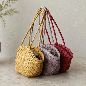Puede incluir: Tres bolsos de mano pequeños y tejidos en forma de semicírculo con un patrón de cestería apretado. Los bolsos son de color amarillo brillante, lavanda suave y rojo intenso, cada uno con dos asas largas trenzadas desde el borde superior.