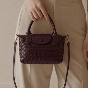 Può includere: Una borsa a mano color bordeaux intenso con un motivo esterno in pelle intrecciata, un manico superiore e una patta con chiusura a bottone a pressione centrale. La borsa include una tracolla sottile rimovibile con clip in metallo dorato.