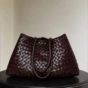 Può includere: Una borsa tote marrone scuro caratterizzata da un intricato motivo intrecciato su tutto l'esterno. La borsa ha un'ampia forma trapezoidale con due manici intrecciati abbinati fissati sulla parte superiore, creando un aspetto uniforme e strutturato.