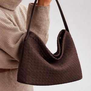 Può includere: Una borsa a tracolla in pelle intrecciata marrone scuro con una lunga tracolla. La borsa ha una forma morbida e ampia e una trama intrecciata. La tracolla è di una tonalità più scura di marrone. La borsa è tenuta da una persona che indossa un maglione beige.