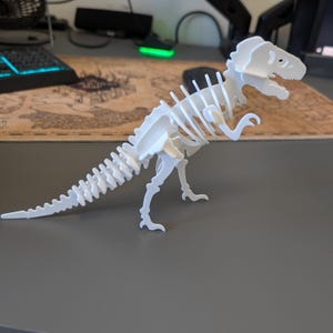 Puede incluir: Un modelo de esqueleto de Tyrannosaurus Rex blanco impreso en 3D. El modelo de dinosaurio está ensamblado, mostrando la caja torácica, la columna vertebral y el cráneo. El modelo está sobre una superficie gris.
