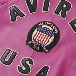 Pode incluir: Close-up de uma jaqueta de couro rosa com letras pretas e brancas que soletram "AVIR" e "USA". Um patch circular apresenta a bandeira americana e as palavras "ALL AMERICANS ESTABLISHED 1975 N.Y."