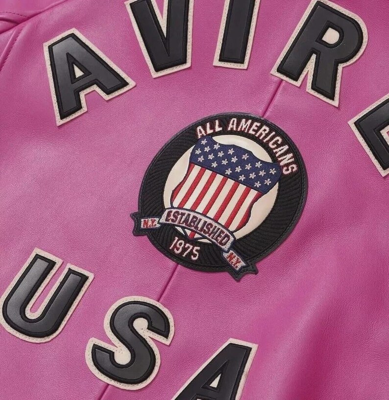 Pode incluir: Close-up de uma jaqueta de couro rosa com letras pretas e brancas que soletram "AVIR" e "USA". Um patch circular apresenta a bandeira americana e as palavras "ALL AMERICANS ESTABLISHED 1975 N.Y."